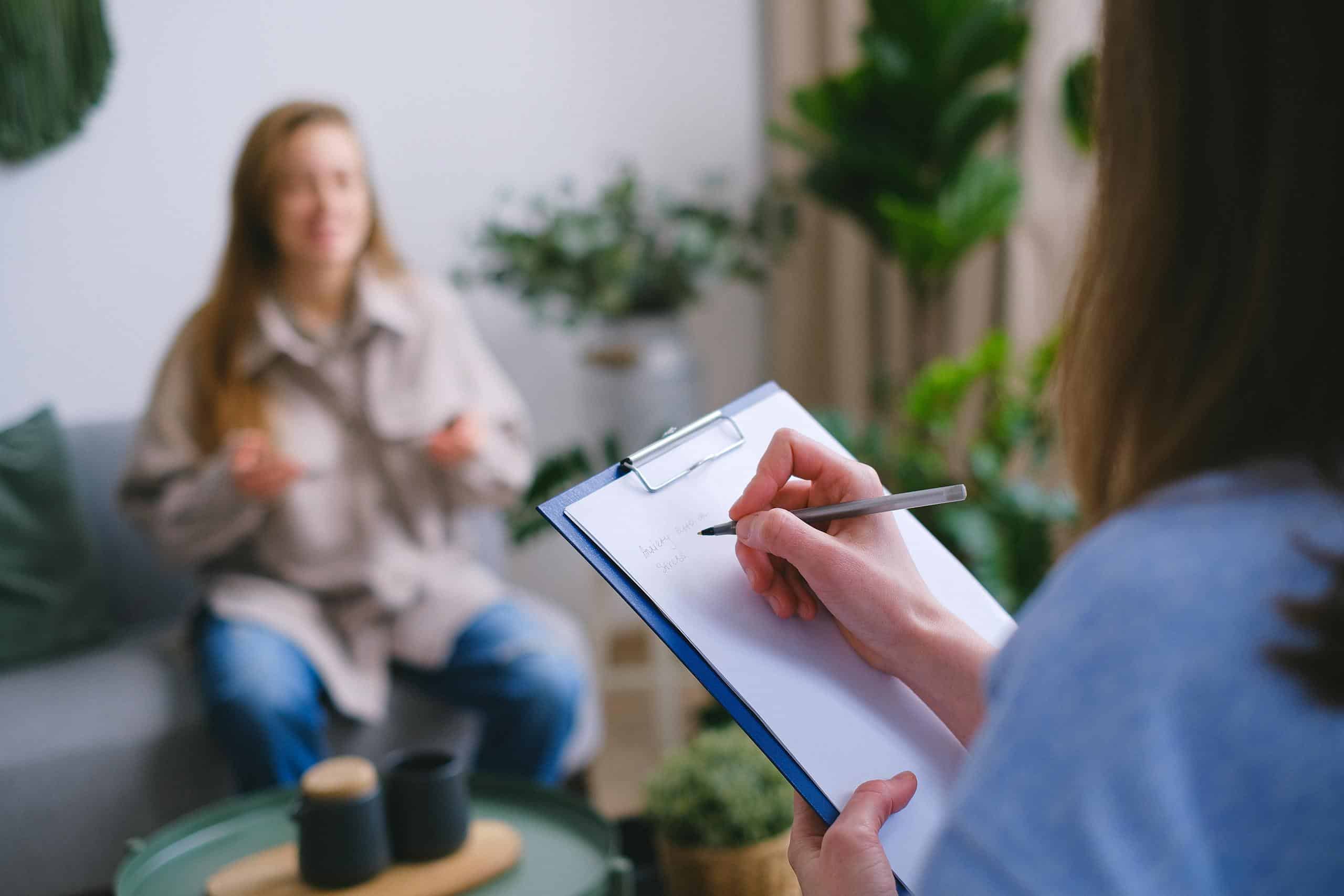 Professional female psychologist writing on clipboard while sitting against client on blurred background during psychotherapy session in light office
