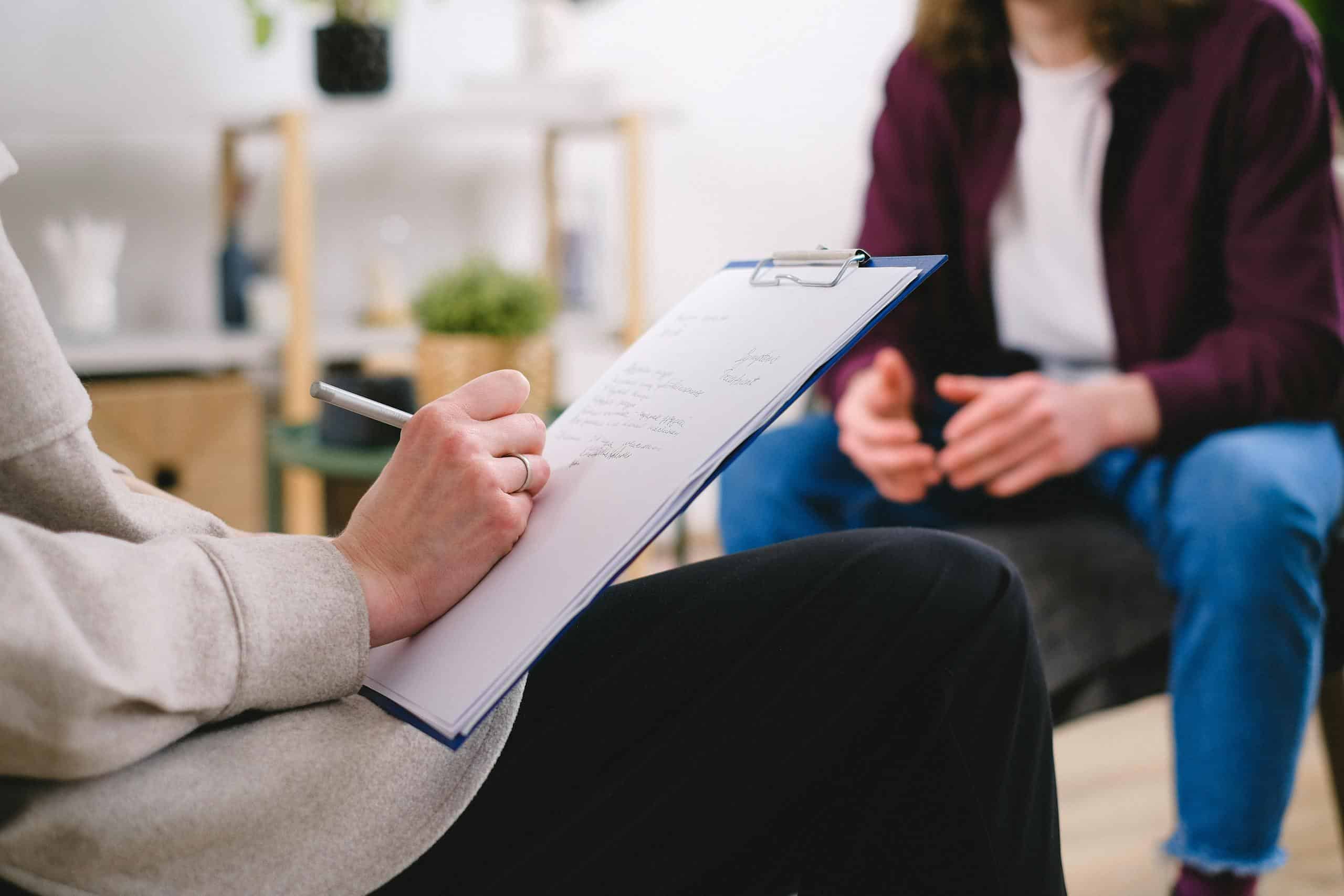 Close-up of a therapist writing notes on a clipboard while conversing with a patient.