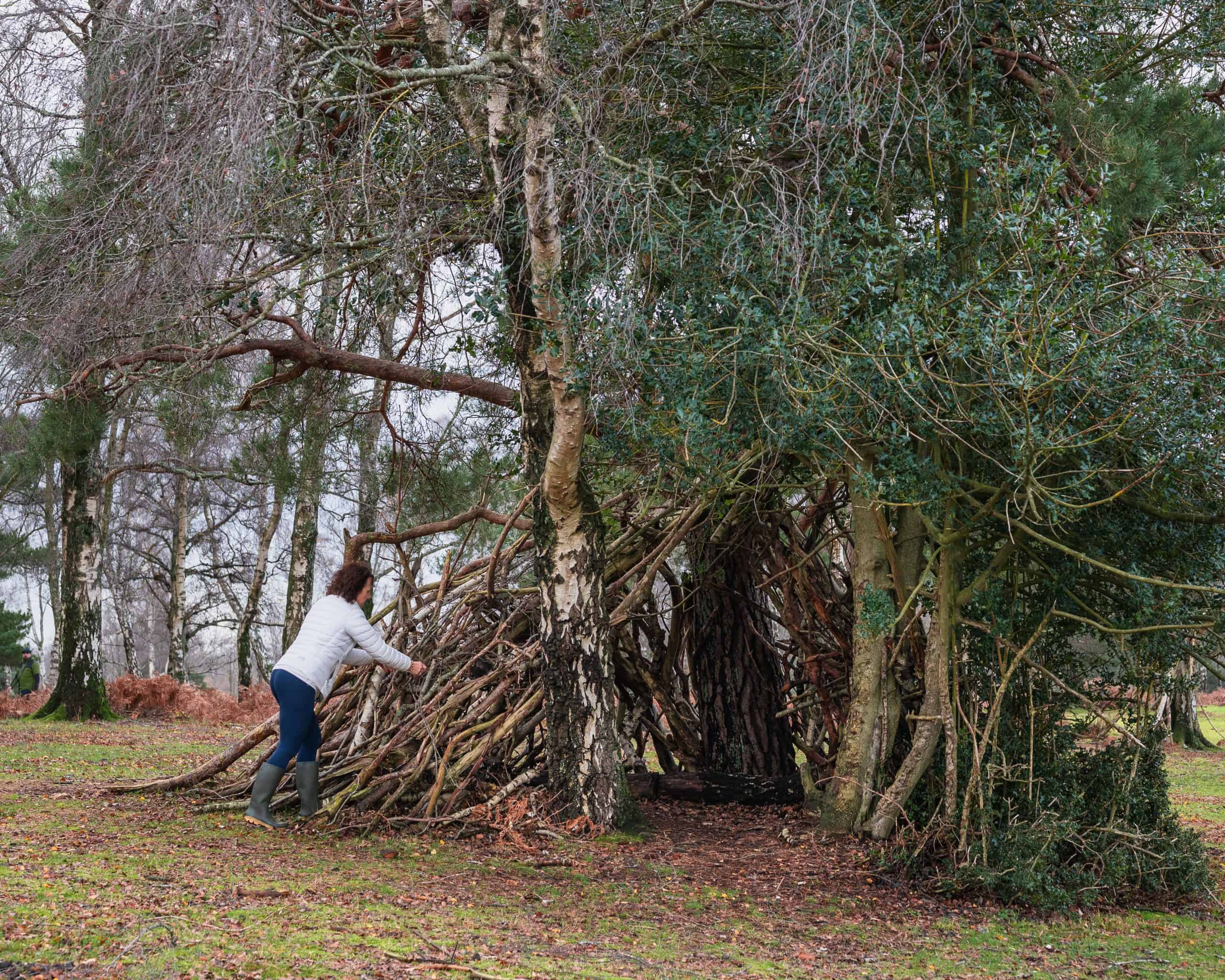 Rebecca building a wooden den with branches under trees in a park.